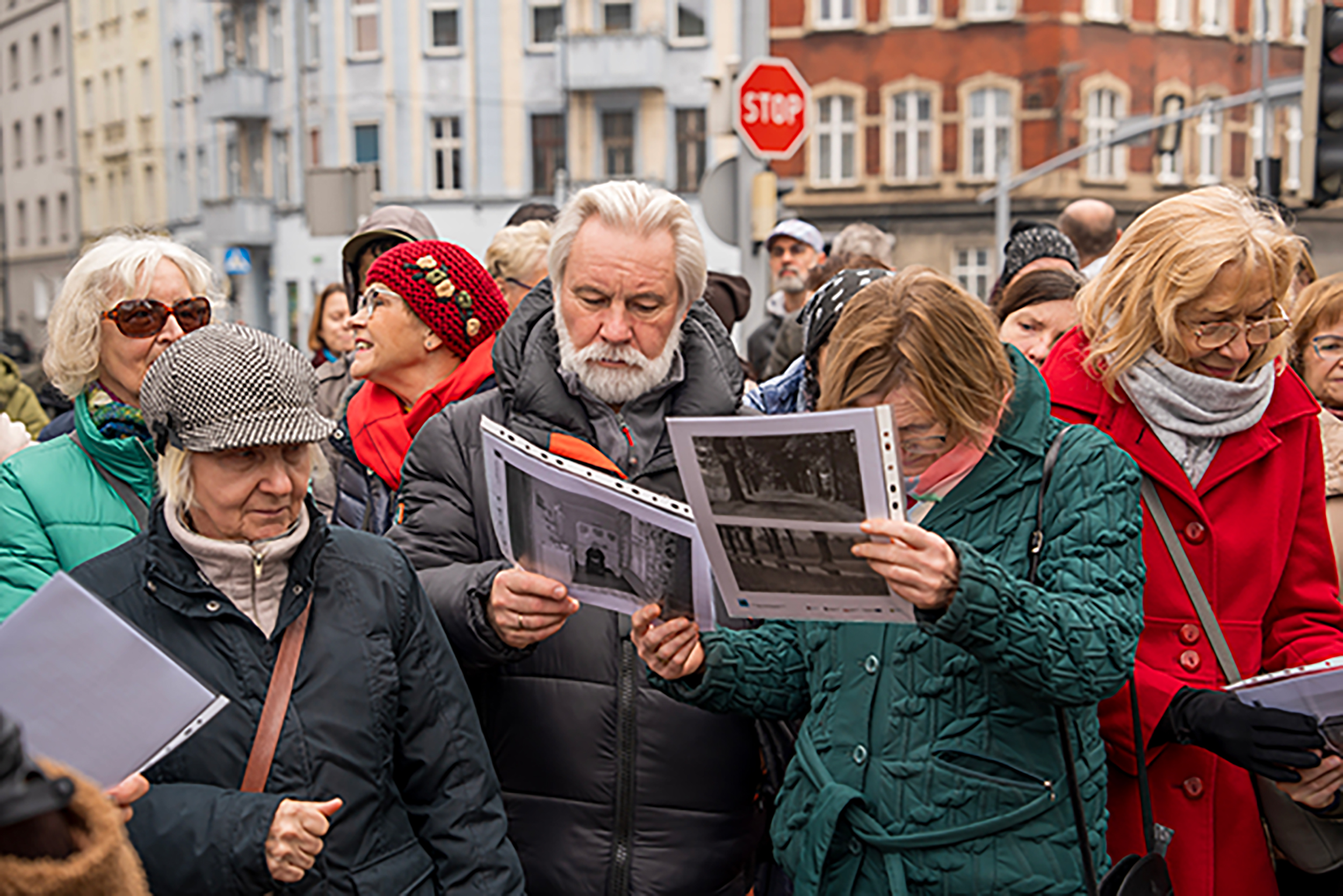 Ludzie biorący udział w spacerze historycznym i przeglądają zdjęcia wydrukowane na kartkach przy ulicy Zwycięstwa w Gliwicach