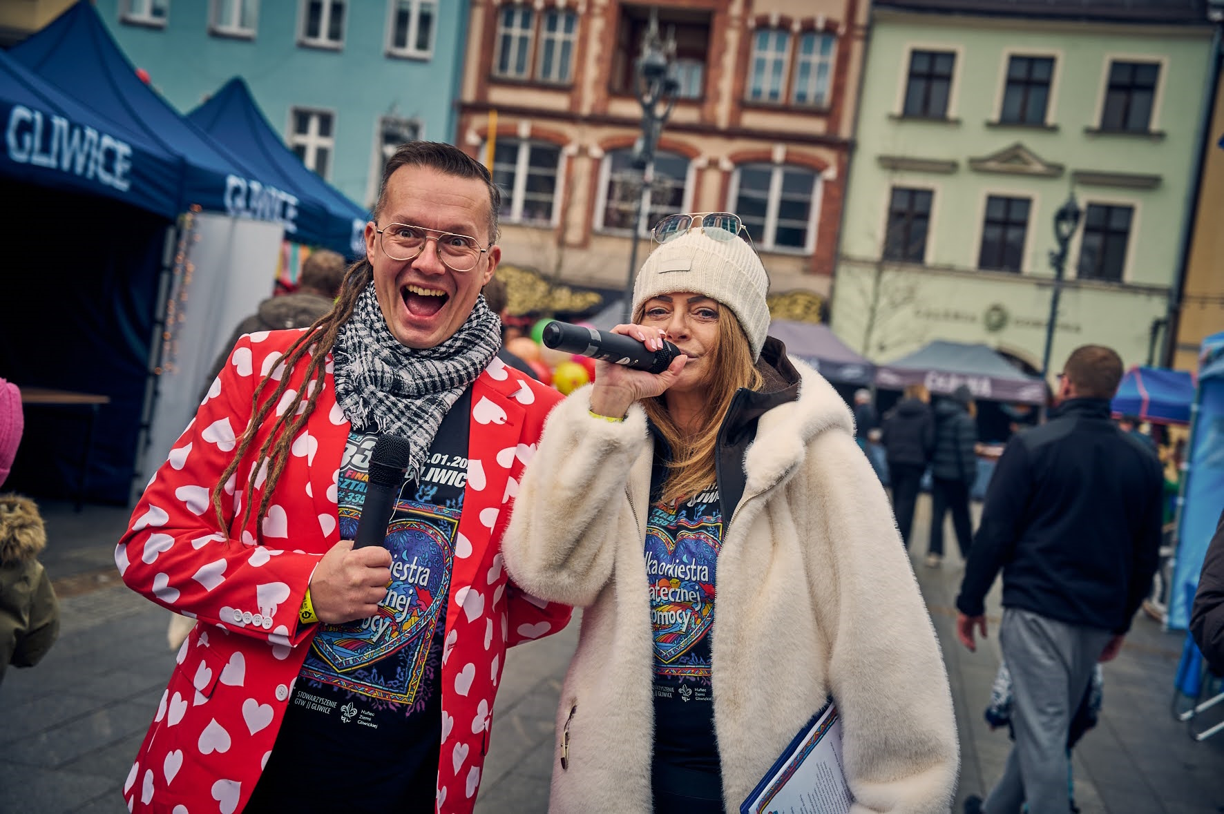 Uśmiechnięty mężczyzna w czerwonej marynarce z białymi serduszkami, obok niego kobieta z mikrofonem