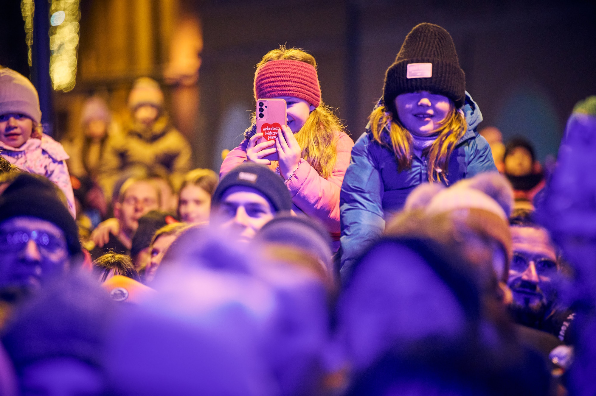 Dziewczynki na ramionach dorosłych ogladają koncert, jedna trzyma telefon, druga się uśmiecha