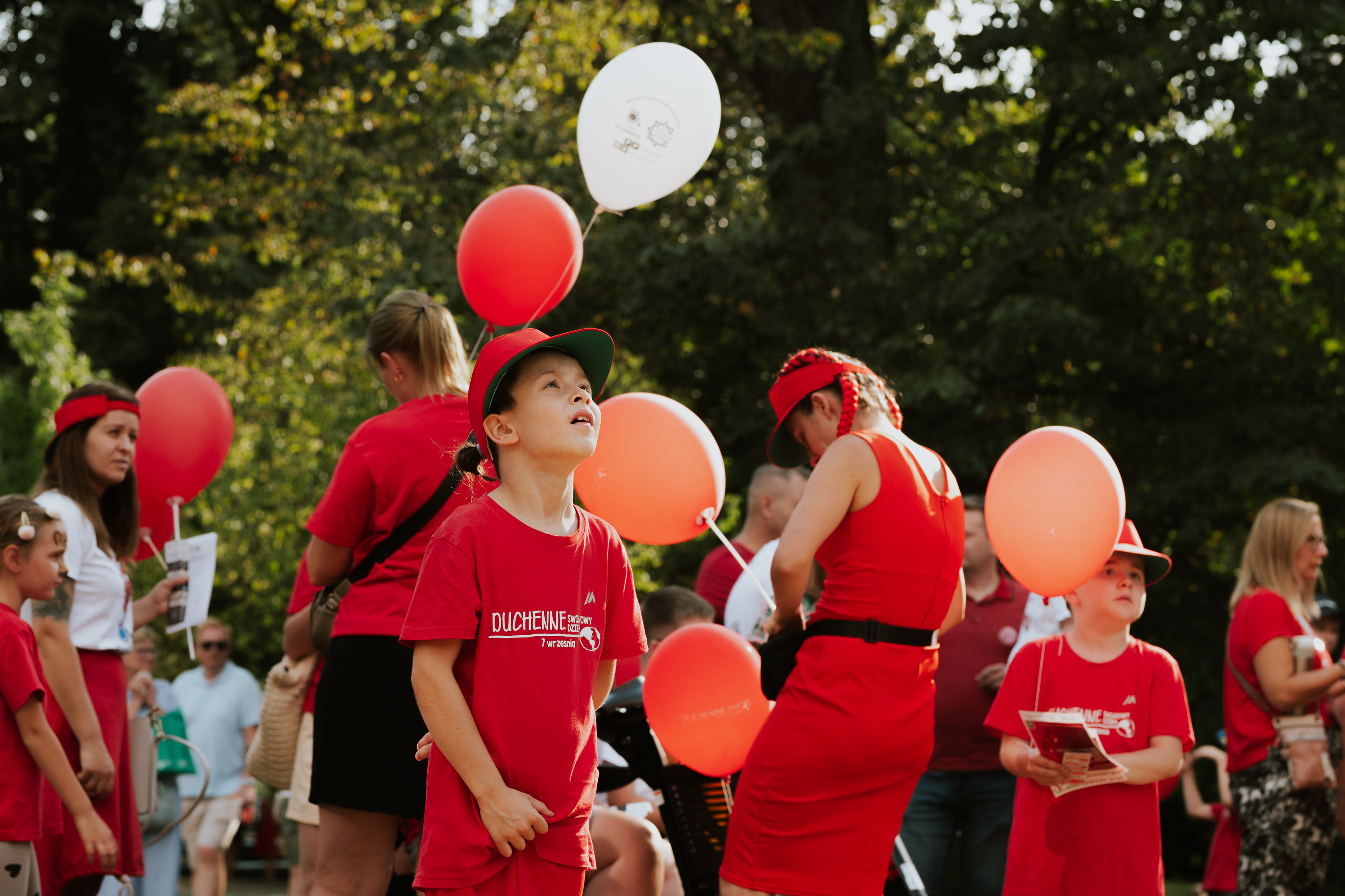 Chłopiec w czerwonej koszulce patrzący w niebo, a za nim czerwone balony