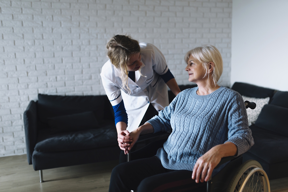 Młoda kobieta w stroju medycznym nachyla się nad starszą kobietą w niebieskim swetrze, siedzącą na wózku. W tle wnętrze pokoju o białych ścianach z dużą ciemną kanapą.