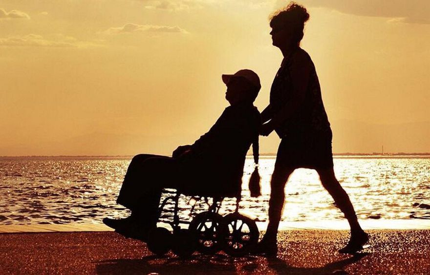 kobieta pcha po plaży wózek iwalidzki z siedzącym mężczyzną